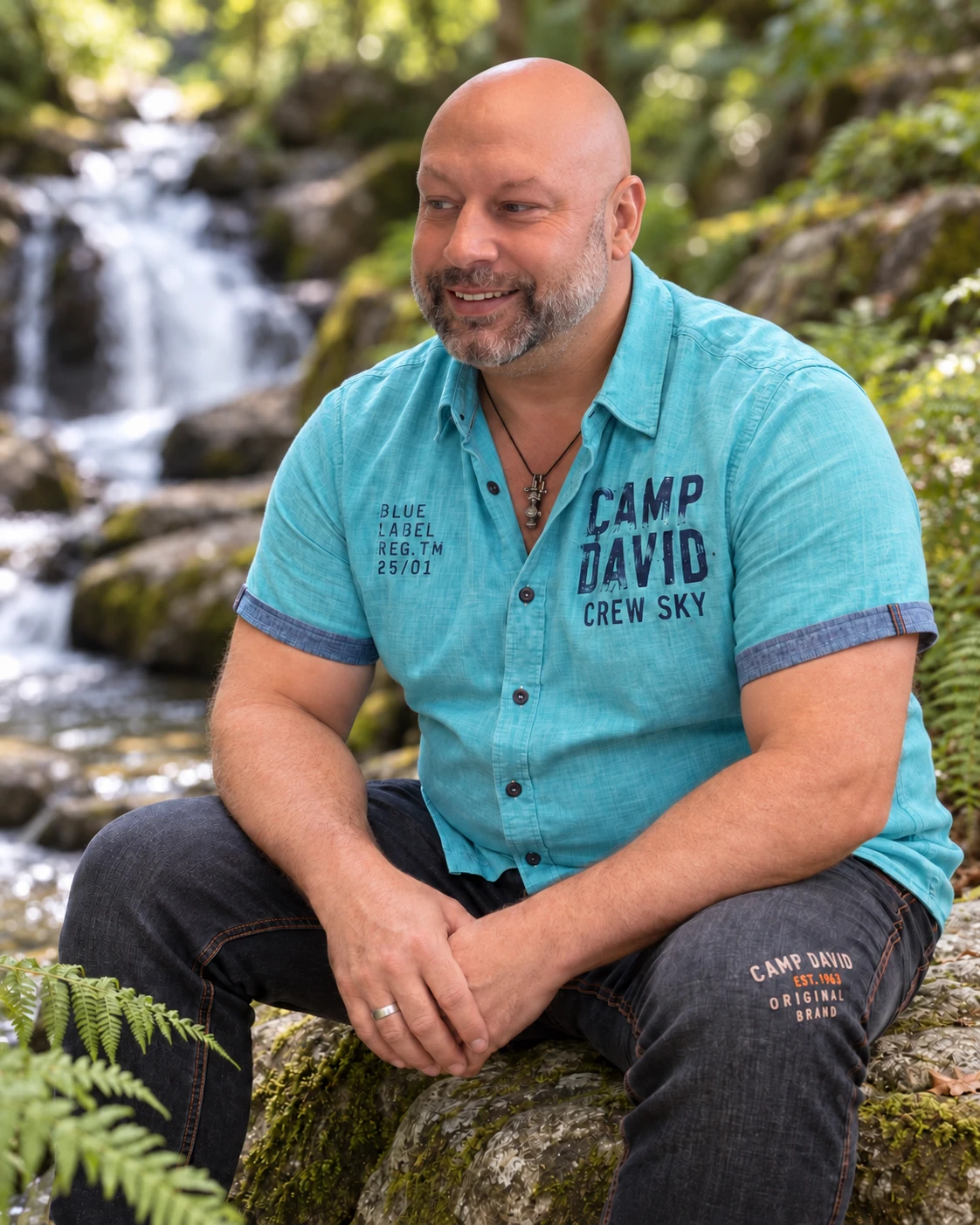 Heiko Gärtner am Wasserfall mit offenem türkisem Camp-David-Hemd, dunkler Jeans und silbernem Ring an der rechten Hand